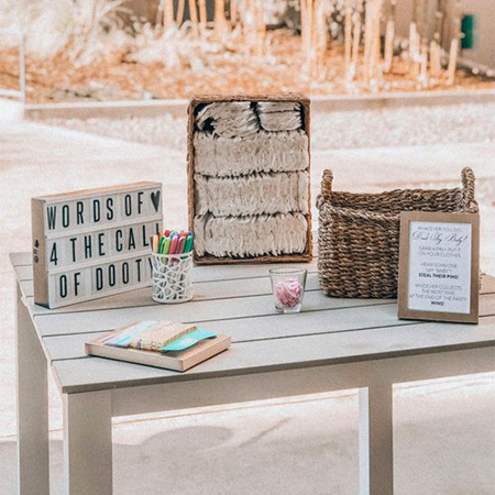 A white outdoor table holds items for a baby shower game, including a light-up sign that reads "WORDS OF 4 THE CALL OF DOOTY," colorful markers, a basket of diapers, and a framed sign with instructions.