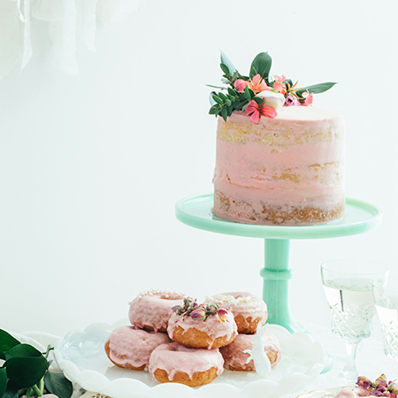 A light pink frosted cake topped with fresh flowers sits on a mint green cake stand, next to a white platter with several pink-glazed donuts decorated with dried flower petals.
