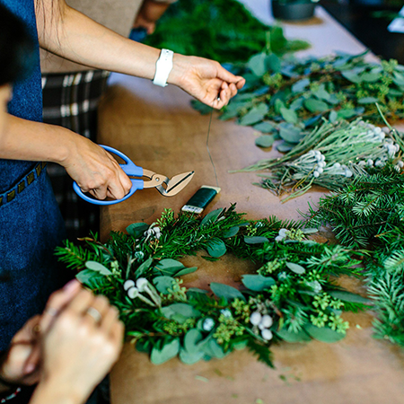A close-up shot shows two people engaged in making a wreath with various green foliage and white berries on a craft table.